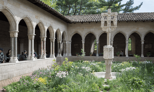 A stone cloister garden with a central ornate cross-shaped fountain, surrounded by blooming flowers and arches, evokes the charm of the Holiday Train Show at Grand Central. People stroll along the shaded arched walkway under a tiled roof, with tall trees in the background.